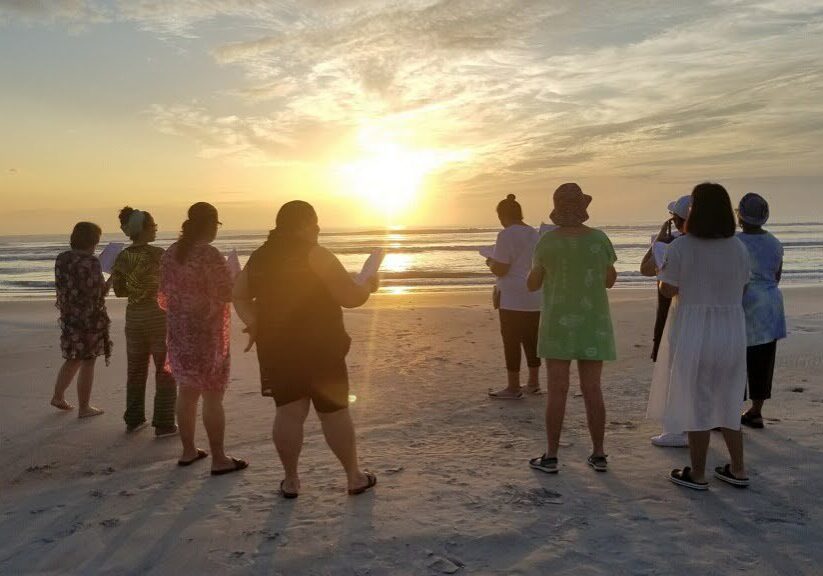 A group of people standing on the beach at sunset.